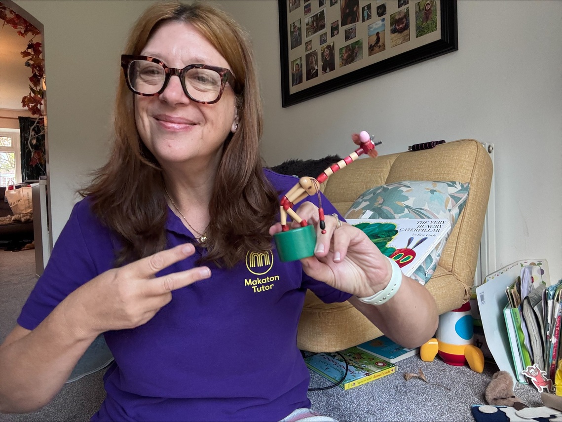 Woman in purple Makaton Tutor top uses makaton sign for again whilst holding a pop up toy