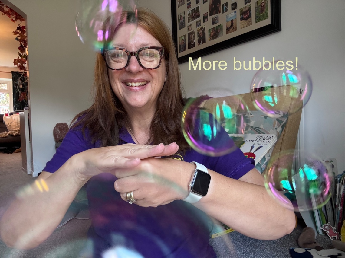 Woman in a purple Makaton Tutor Top uses the makaton sign for more whilst bubbles are blowing 